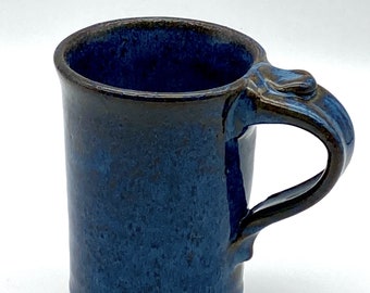 May include: A blue ceramic mug with a brown rim and handle. The mug has a speckled glaze and a unique, slightly curved handle.