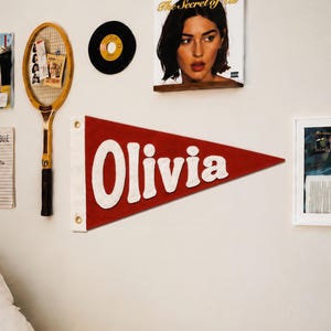 May include: A triangular red felt pennant featuring the name Olivia in large white felt letters. The pennant has a white vertical strip on the left side with two brass-coloured metal grommets for hanging, creating a classic sports-style banner.