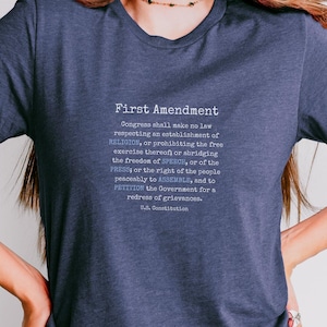 May include: A heather navy blue short-sleeve t-shirt featuring the text of the First Amendment of the U.S. Constitution printed in white. The graphic includes the title First Amendment followed by the full text and U.S. Constitution at the bottom.