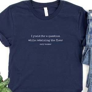 May include: A navy blue t-shirt with the text 'I yield for a question while retaining the floor cory booker' printed on the front.
