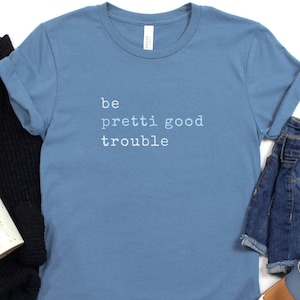 May include: A steel blue t-shirt with the words "be pretti good trouble" printed in white. The shirt is styled with denim shorts, brown sandals, a black sweater, and a white mug with black speckles.