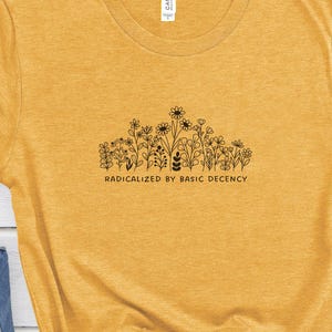 Könnte beinhalten: Ein senfgelbes T-Shirt mit einer schwarzen Strichzeichnung verschiedener Wildblumen und dem mittig aufgedruckten Text "RADICALIZED BY BASIC DECENCY". Das T-Shirt wird zusammen mit einer blauen Jeans, einer schwarzen Sonnenbrille und schwarzen Turnschuhen gezeigt.