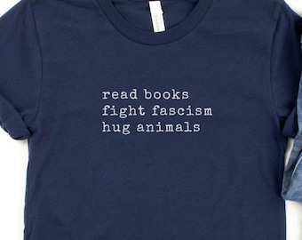 Könnte beinhalten: Marineblaues T-Shirt mit dem weißen Text "read books, fight fascism, hug animals". Das Shirt wird mit einer blauen Jeans und einer kleinen grünen Topfpflanze präsentiert.