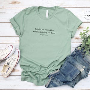 May include: A sage green t-shirt with the text 'I yield for a question while retaining the floor' and 'cory booker' printed on the front. The t-shirt is laying on a white wooden surface with a pair of white converse shoes and a pair of blue jeans.