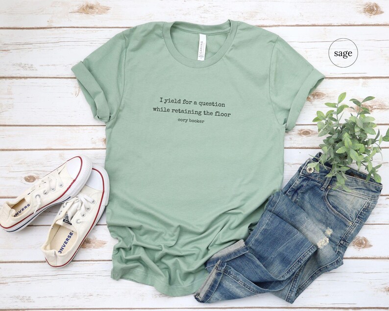 May include: A sage green t-shirt with the text 'I yield for a question while retaining the floor' and 'cory booker' printed on the front. The t-shirt is laying on a white wooden surface with a pair of white converse shoes and a pair of blue jeans.
