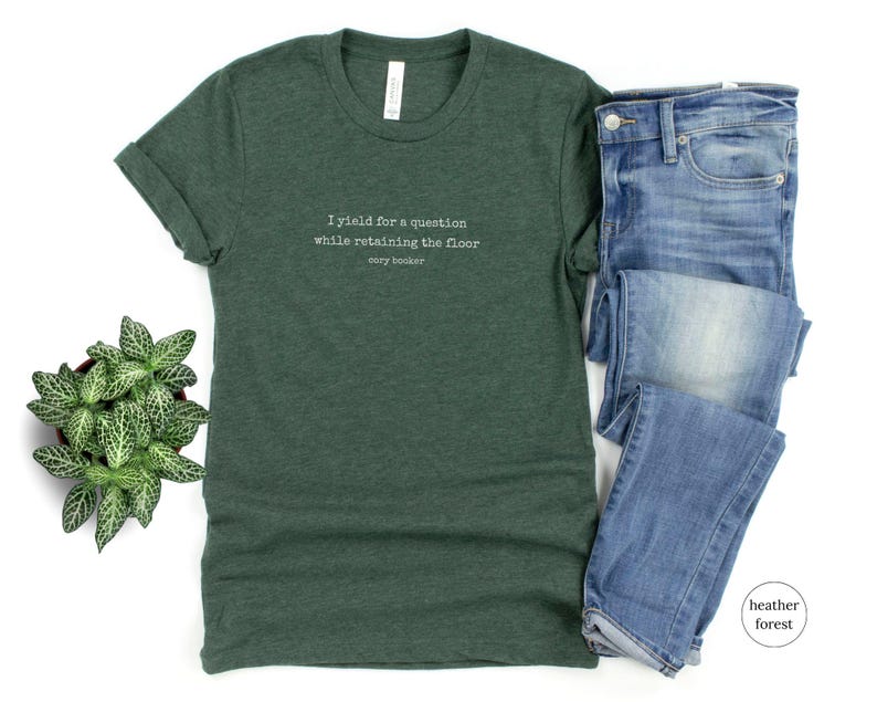 May include: A green t-shirt with the text 'I yield for a question while retaining the floor Cory Booker' printed on the front.
