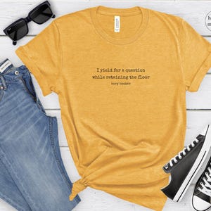 May include: A heather mustard colored t-shirt with the text 'I yield for a question while retaining the floor Cory Booker' printed on the front.