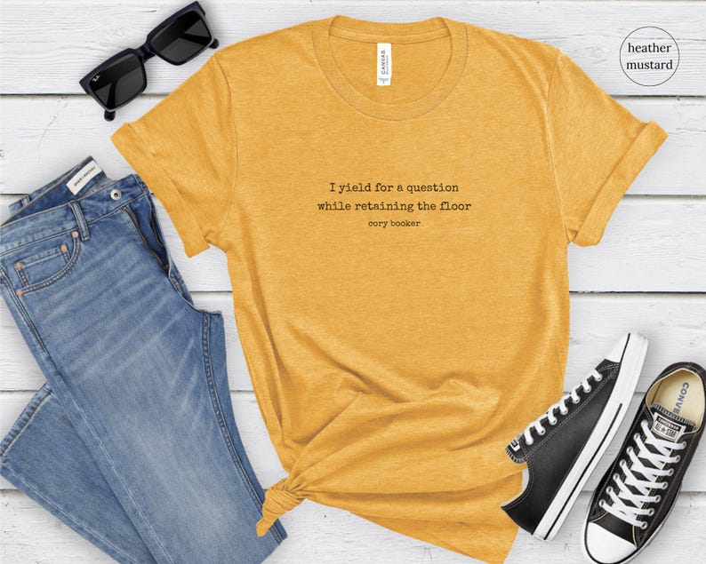 May include: A heather mustard colored t-shirt with the text 'I yield for a question while retaining the floor Cory Booker' printed on the front.