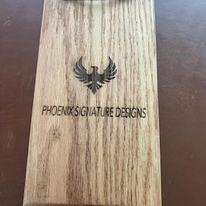 May include: Wooden clipboard with a black metal clip at the top. The wood has a natural grain pattern and features a laser-etched phoenix logo and the text "PHOENIX SIGNATURE DESIGNS".
