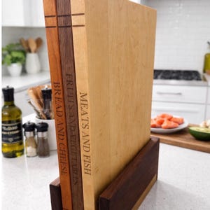 May include: Three wooden chopping boards in a dark wood holder. The boards are labelled "Bread and Cheese", "Fruits and Vegetables", and "Meats and Fish". The boards are different shades of brown and tan.