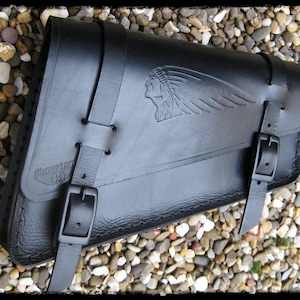 May include: Black leather motorcycle saddlebag with an embossed Indian head logo and a stitched design. The bag has a buckle closure and a strap.