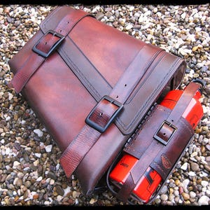 Könnte beinhalten: Eine braune Ledertasche mit zwei Riemen und schwarzen Schnallen. Neben der Tasche befindet sich ein passender Leder-Flaschenhalter mit einer roten Wasserflasche. Auf der Wasserflasche steht das Wort "LAKEN".
