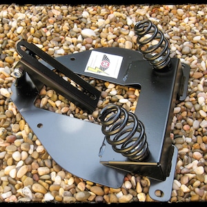 May include: A black metal bracket with two coiled springs and a slotted arm. The bracket has a sticker with a logo and is set against a background of small, multicolored pebbles. The springs are approximately 4 inches tall.