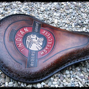 Peut inclure: Selle de moto en cuir marron avec le logo "Indian Motorcycle Riders Group" en rouge et blanc. La selle a une bordure décorative et est posée sur un fond de petits cailloux.