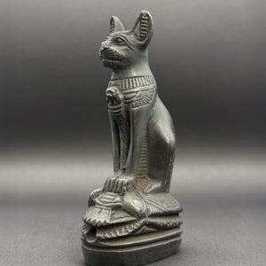 Puede incluir: Una figura de color gris oscuro con acabado mate de un gato sentado en estilo egipcio. El gato tiene orejas puntiagudas, un collar con detalles grabados y se asienta sobre una base rectangular con líneas horizontales y una pequeña abertura.
