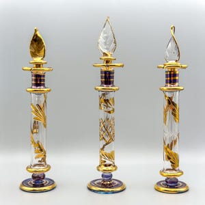 May include: Three decorative glass bottles with gold accents and purple and blue bases. The bottles have different designs on the body and are perfect for displaying perfume or essential oils.