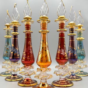 May include: Set of seven decorative glass bottles with gold accents and intricate designs. The bottles have clear glass stoppers shaped like flames. The bottles are arranged in a row on a white surface.