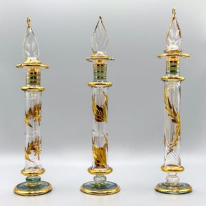 May include: Three clear glass perfume bottles with gold leaf accents and a green and gold base. The bottles have a decorative, ornate design.