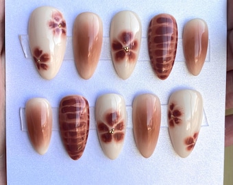May include: Set of ten false nails with different designs. Some nails have a brown ombre effect, others have a brown and white floral design, and others have a brown and white striped design.