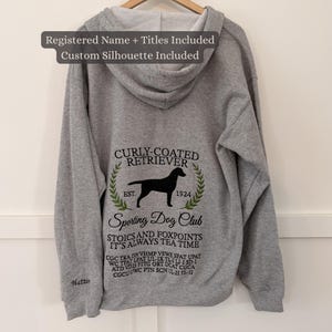 May include: A gray hoodie with a hood and drawstring, featuring a black Curly-Coated Retriever silhouette and text. The design includes the words "Curly-Coated Retriever," "Sporting Dog Club," and other text. The hoodie is hanging on a wooden hanger.