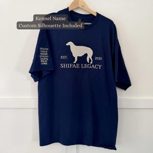 Może przedstawiać: Granatowy t-shirt z krótkim rękawem z białą sylwetką psa, napisem "EST. 2020", "SHIFAE LEGACY" oraz listą imion na rękawie, w tym "SHILOH", "FABLE", "REIGN", "KNIGHT", "DAEVA", "RELIC" i "LORE".