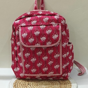 Op de afbeelding: Een gewatteerde rugzak in een levendige magenta tint, versierd met een herhalend wit bloemenpatroon. De rugzak heeft een voorvak, een zijvak en een handvat aan de bovenkant. Witte gestreepte randen omlijnen de randen. De tas staat op een ronde, geweven mat.