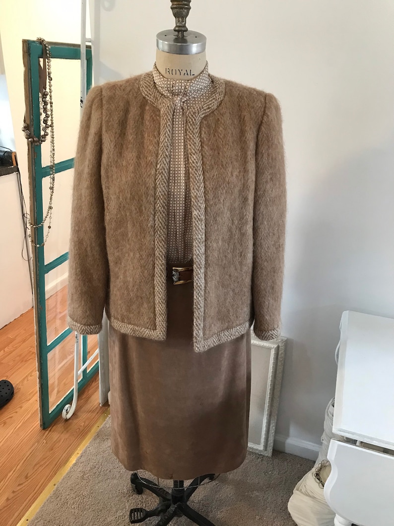 May include: A brown fuzzy jacket with a white and brown trim, worn over a white and brown patterned blouse and a brown skirt.