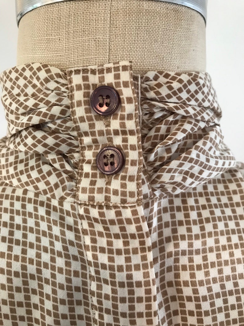 May include: A white blouse with a brown geometric pattern and a ruffled collar. The collar has two brown buttons.