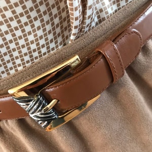 May include: A brown leather belt with a gold and silver buckle. The belt is fastened around a brown fabric garment.