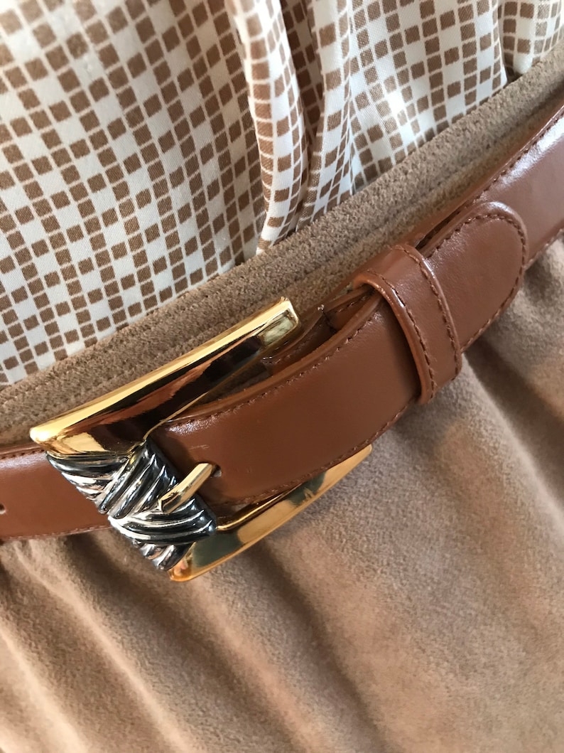 May include: A brown leather belt with a gold and silver buckle. The belt is fastened around a brown fabric garment.