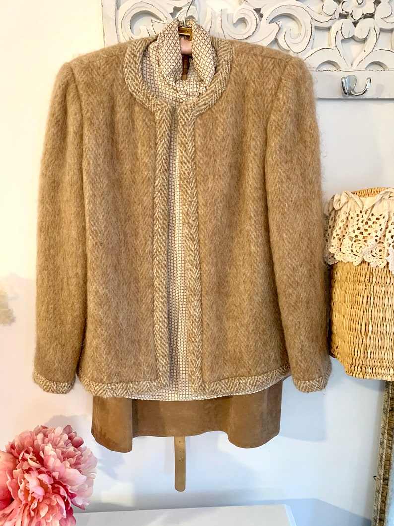 May include: A brown fuzzy jacket with a herringbone pattern and white trim. The jacket is hanging on a hanger and has a white and brown patterned shirt underneath.