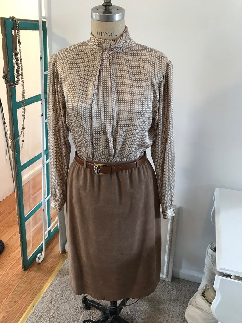 May include: A brown suede skirt with a brown leather belt and a white and brown patterned blouse with a high collar.