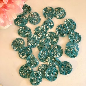 May include: A collection of teal fabric rosettes with a white floral pattern. The rosettes are arranged in a random pattern on a white surface.