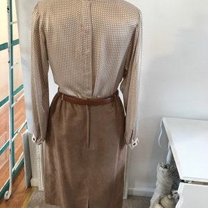 May include: A brown suede skirt with a brown leather belt and a white and brown patterned blouse. The blouse has a high collar and long sleeves.