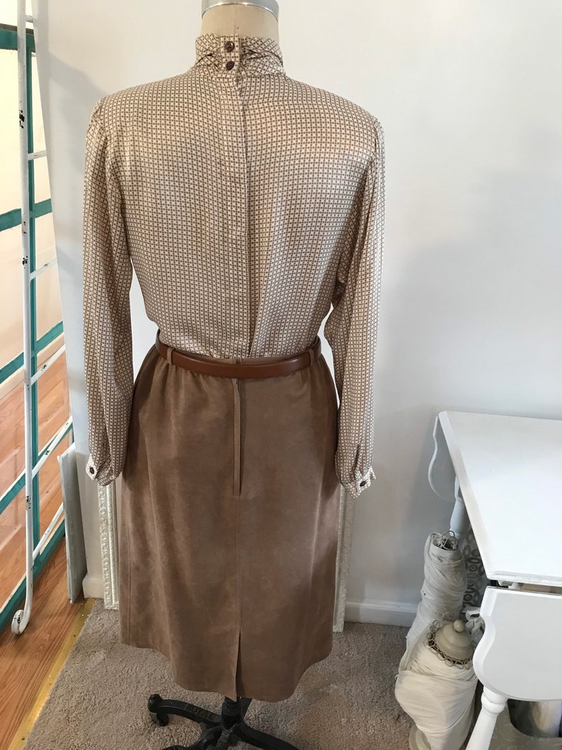 May include: A brown suede skirt with a brown leather belt and a white and brown patterned blouse. The blouse has a high collar and long sleeves.