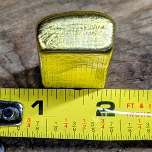 May include: A gold-colored, rectangular object with a textured surface, approximately 1.5 inches long, is positioned next to a yellow measuring tape. The tape shows measurements in inches and fractions of an inch.