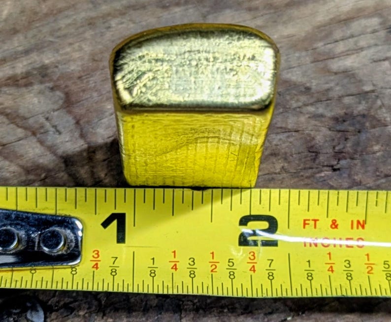 May include: A gold-colored, rectangular object with a textured surface, approximately 1.5 inches long, is positioned next to a yellow measuring tape. The tape shows measurements in inches and fractions of an inch.