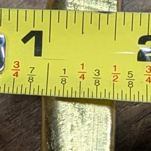 May include: A gold-colored rectangular bar is measured with a yellow tape measure. The tape measure shows the bar is approximately 1 inch wide. The bar has a textured surface.