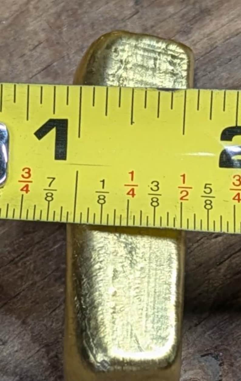 May include: A gold-colored rectangular bar is measured with a yellow tape measure. The tape measure shows the bar is approximately 1 inch wide. The bar has a textured surface.