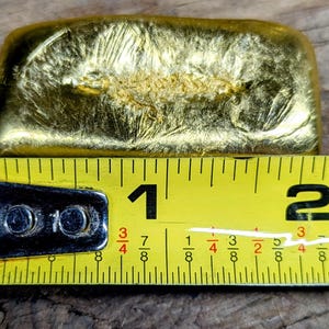 May include: A gold-colored rectangular bar with a textured surface, approximately 1 inch long, is measured with a yellow tape measure. The tape measure has black markings and numbers. The bar is on a wooden surface.