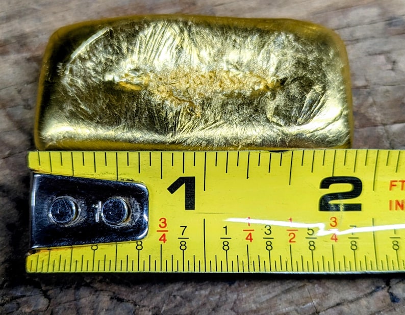 May include: A gold-colored rectangular bar with a textured surface, approximately 1 inch long, is measured with a yellow tape measure. The tape measure has black markings and numbers. The bar is on a wooden surface.