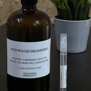 May include: A brown glass bottle with a white label that reads "OUD WOOD DRAMMING". The label also includes the text "CAUTION: FLAMMABLE UNTIL DRY DO NOT USE NEAR FIRE; FLAME OR HEAT 1 Litre (33.814 FL.OZ) TOEJ500000 9999204571". A clear glass spray bottle with a white label that reads "TOM FORD OUD WOOD" is also pictured.
