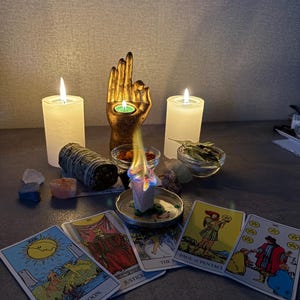 Puede incluir: Una escultura de mano dorada que sostiene una vela de t&eacute;, flanqueada por dos velas de pilar blancas. Un bloque de incienso encendido descansa en un plato de metal, rodeado de cartas del tarot con los textos The Moon, Justice, Page of Pentacles y Six of Pentacles.