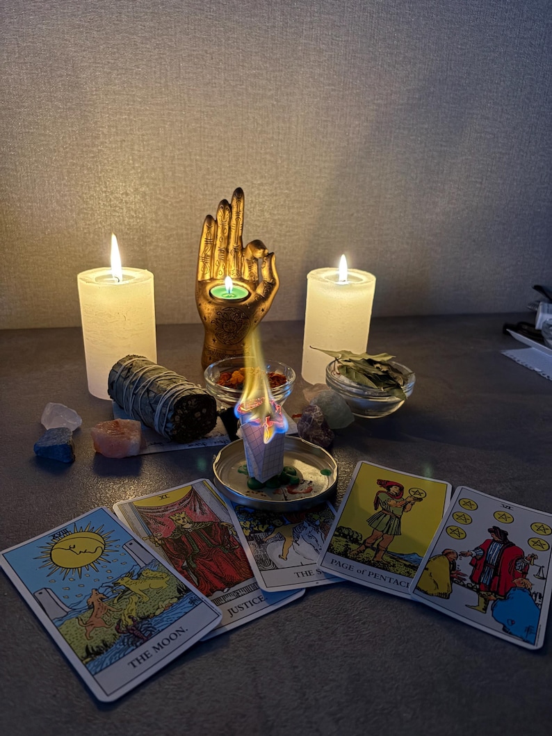 Puede incluir: Una escultura de mano dorada que sostiene una vela de t&eacute;, flanqueada por dos velas de pilar blancas. Un bloque de incienso encendido descansa en un plato de metal, rodeado de cartas del tarot con los textos The Moon, Justice, Page of Pentacles y Six of Pentacles.