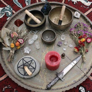 May include: A collection of spiritual items arranged on a decorative metal tray. The items include singing bowls, crystals, a candle, a ritual knife, dried flowers, and a stone with a pentagram symbol. The tray is set on a patterned red rug.