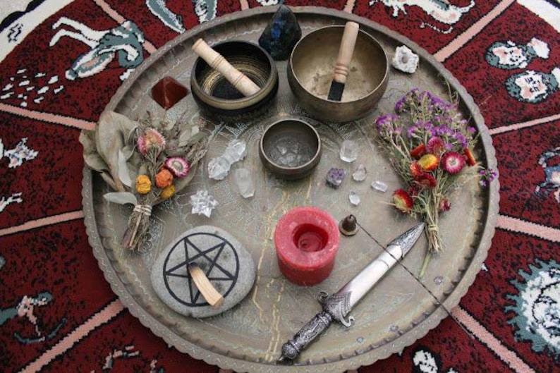 May include: A collection of spiritual items arranged on a decorative metal tray. The items include singing bowls, crystals, a candle, a ritual knife, dried flowers, and a stone with a pentagram symbol. The tray is set on a patterned red rug.