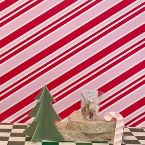 Könnte beinhalten: Eine festliche Weihnachtsdekoration mit einem grünen Papiertannenbaum, einer Zuckerstange und einem kleinen Ornament in einer transparenten Verpackung. Der Hintergrund hat ein rot-rosa Streifenmuster, und die Basis ist ein schwarz-weißes Schachbrettmuster.