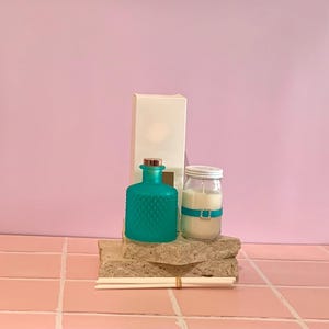 May include: A teal glass diffuser bottle with a diamond-patterned texture, a white candle in a glass jar with a teal ribbon and rhinestone buckle, a tall white rectangular box, and a bundle of white reed diffuser sticks arranged together.