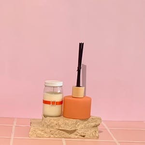 May include: A candle in a glass jar with a white lid and an orange ribbon with a silver buckle. Beside it is an orange diffuser with black reeds and a wooden top. The items are on a stone block against a pink backdrop.