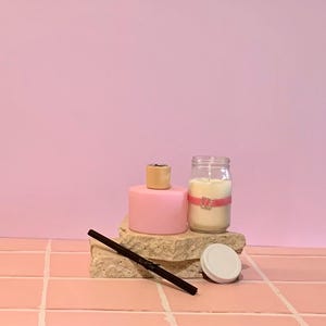 May include: A pink candle, a pink diffuser with a wooden top, and a candle in a jar with a pink ribbon are displayed on a stone. A black stick and a white lid are also present. The background is a solid pink colour.